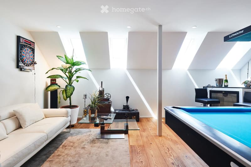 Modern, well-lit room with a white sofa, glass coffee table, bar area with stools, dartboard on the wall, and a large potted plant.
