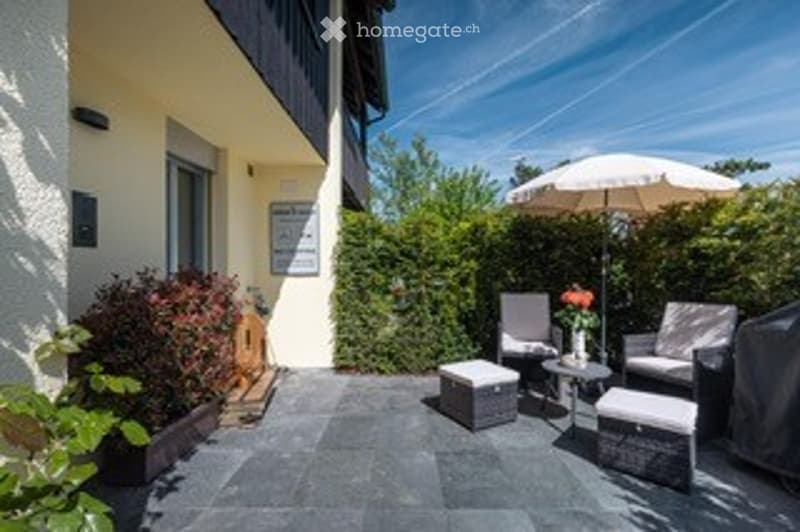 A small patio area with a table, chairs, and an umbrella, surrounded by greenery and a building entrance.