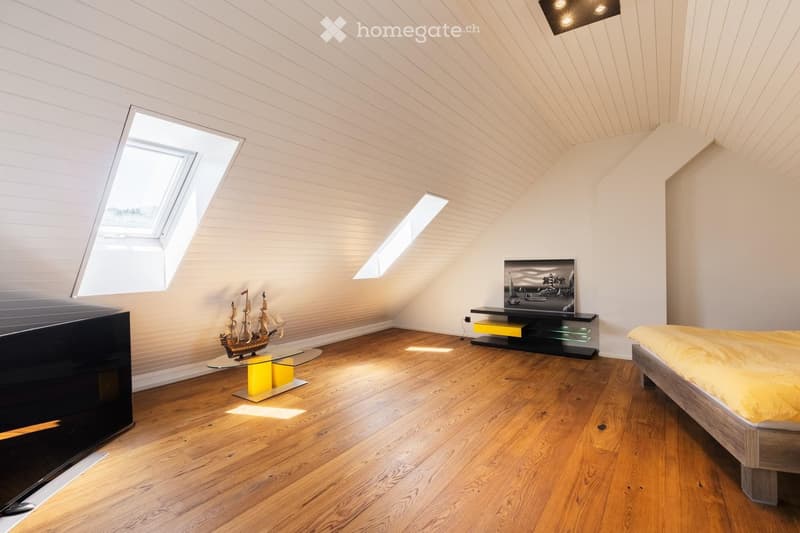 Modern bedroom with wooden flooring, slanted ceiling, skylight windows, a bed with yellow bedding, a glass table with a model ship, and a TV stand with a mounted TV.