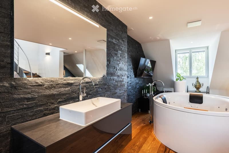 Modern bathroom with a stone accent wall, large sink, bathtub, mirror, and television mounted on the wall. Features wooden flooring and a sleek design.