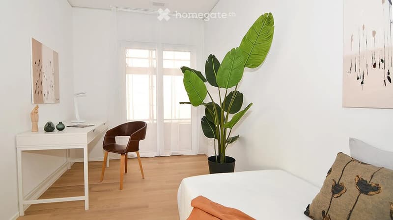 Modern bedroom with a white bed, a brown leather chair, a white desk with a lamp, and a large leafy plant in a black pot. The room features light wooden flooring and white walls with abstract artwork.