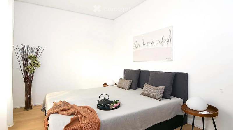 Modern bedroom with a neatly made bed, decorative pillows, a small table with a teapot and cups, a vase with dried branches, and a piece of abstract artwork on the wall.