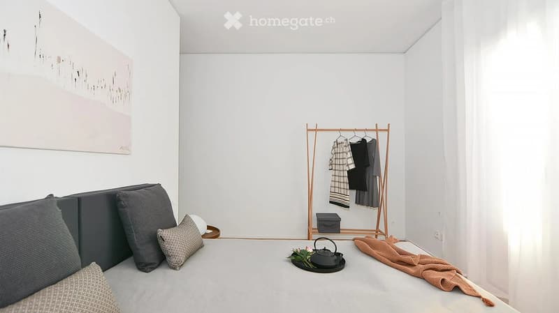 A modern, minimalist living space with a grey sofa, decorative pillows, a small teapot with flowers, a clothing rack with folded clothes, and a folded blanket on the floor.