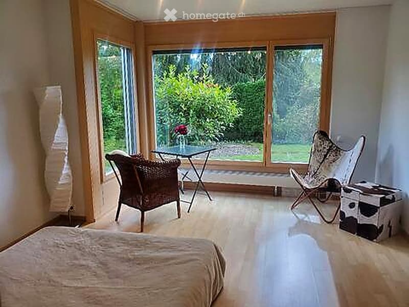 Bright, modern bedroom with large windows overlooking a garden, featuring a bed, wicker chair, small table, and decorative stool.
