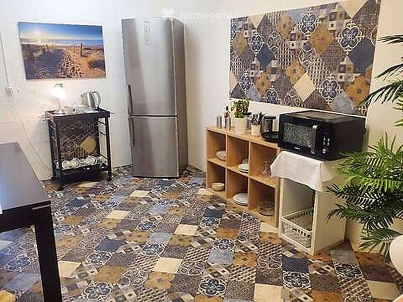 A small, furnished room with a refrigerator, microwave, small table, chair, lamp, potted plant, and picture frame on the wall.