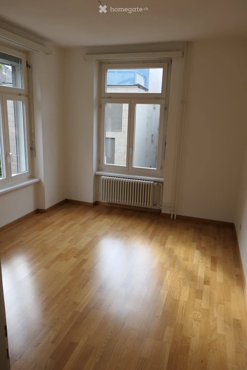 Empty room with wooden flooring, white walls, and large windows with blinds. A radiator is visible beneath one window.