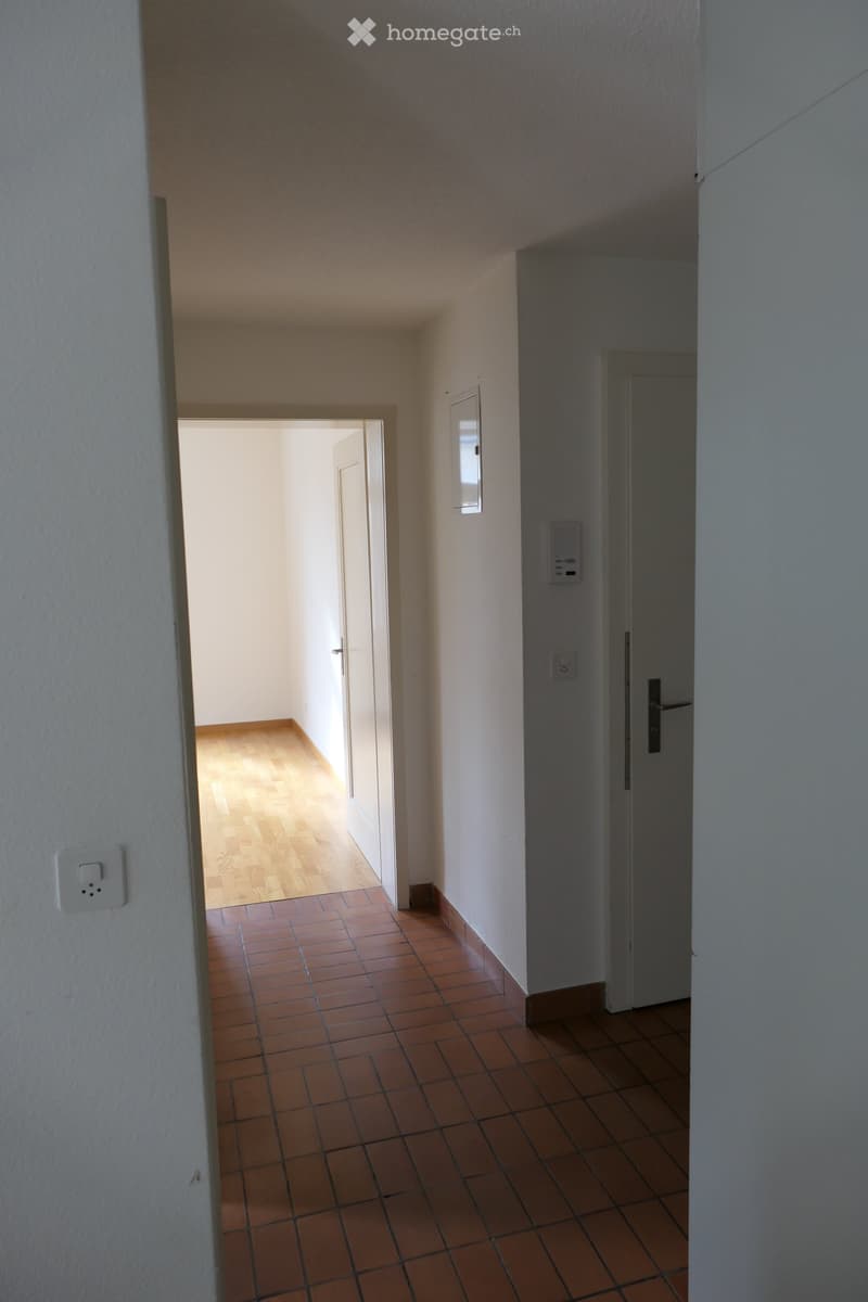 Empty hallway with tiled floor and wooden flooring visible in the adjacent room. White walls and a closed door on the right side.
