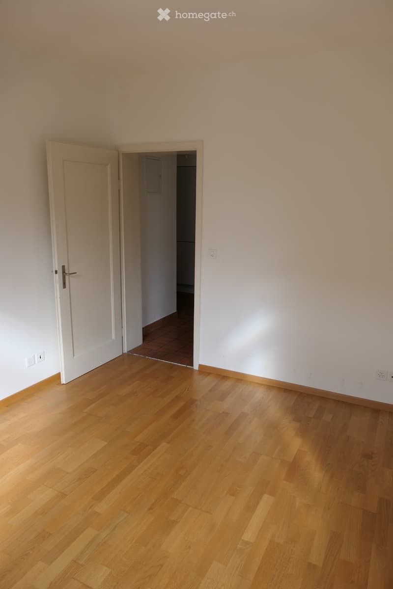 Empty room with light wooden flooring, white walls, and an open door leading to another room with tiled flooring.