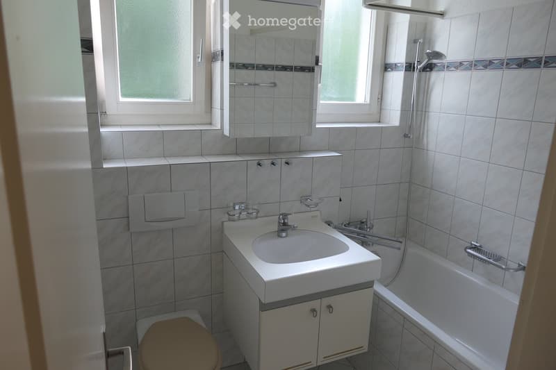Modern bathroom with white tiles, a sink with a faucet, a toilet, a bathtub with a showerhead, and a mirror above the sink.