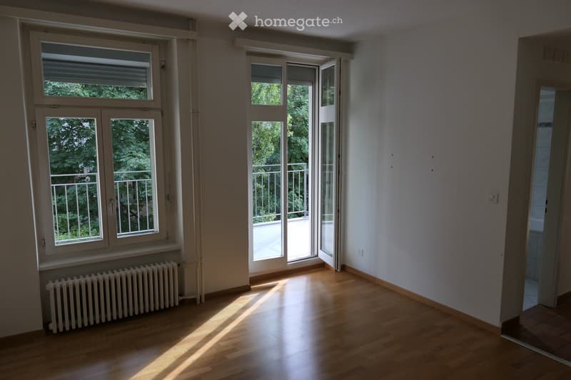 Empty room with wooden flooring, large windows, and a radiator. The room has a balcony accessible through a sliding glass door.