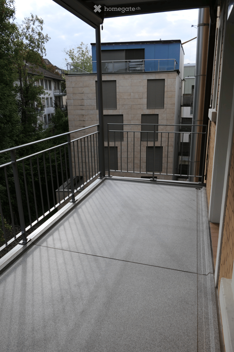 Modern balcony with metal railing, concrete floor, and view of neighboring buildings and greenery.