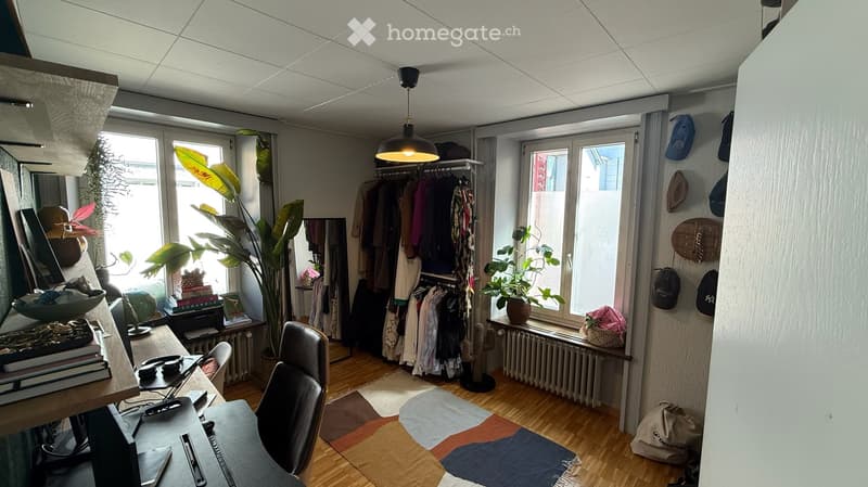 A well-lit room with wooden flooring, a large window, a desk with a laptop, a chair, a cat tree, and several plants. There is a clothing rack with hanging clothes, a mirror, and various decorative items on the walls and surfaces.
