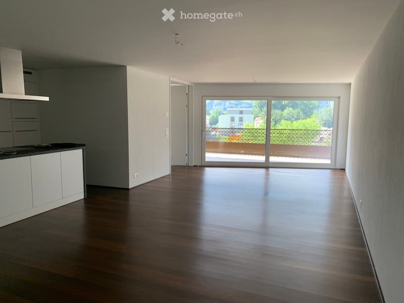 Empty room with wooden flooring, white walls, and a sliding glass door leading to a balcony with a view of greenery and a building.