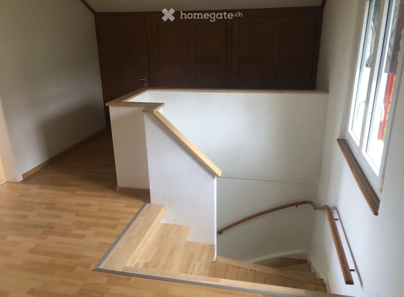 Spacious hallway with wooden flooring, white walls, and a staircase leading upwards. The staircase features wooden railings and steps, with a window on the right side allowing natural light to enter.