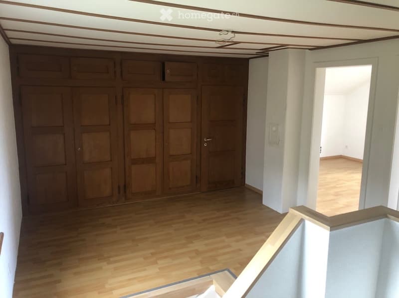 Empty hallway with wooden flooring, white walls, and a large wooden wardrobe on one side. The hallway leads to another room with similar flooring.
