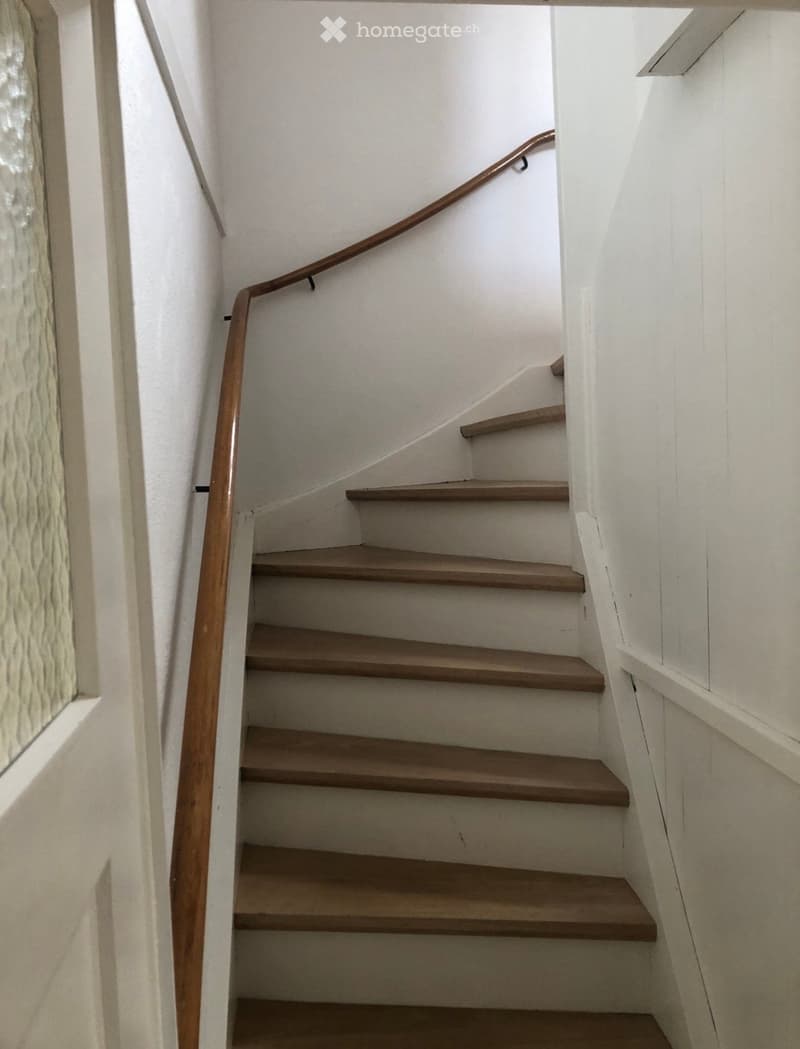 Wooden staircase with a wooden handrail, white walls, and a textured wall panel on the left side.
