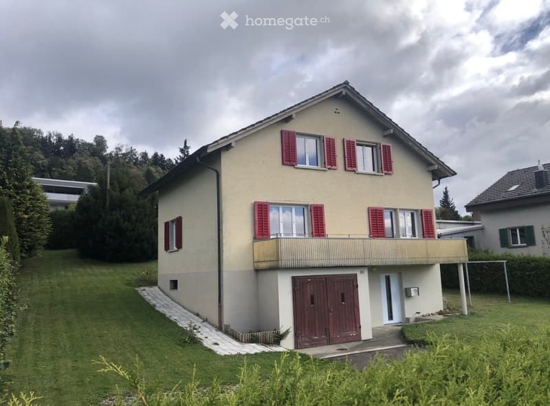 Two-story house with beige walls, red shutters, and a balcony on the upper floor. Features a garage entrance and a well-maintained lawn.