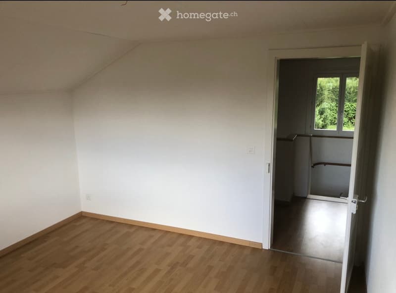 Empty room with light wooden flooring and white walls, featuring an open doorway leading to another room with a window showing greenery outside.