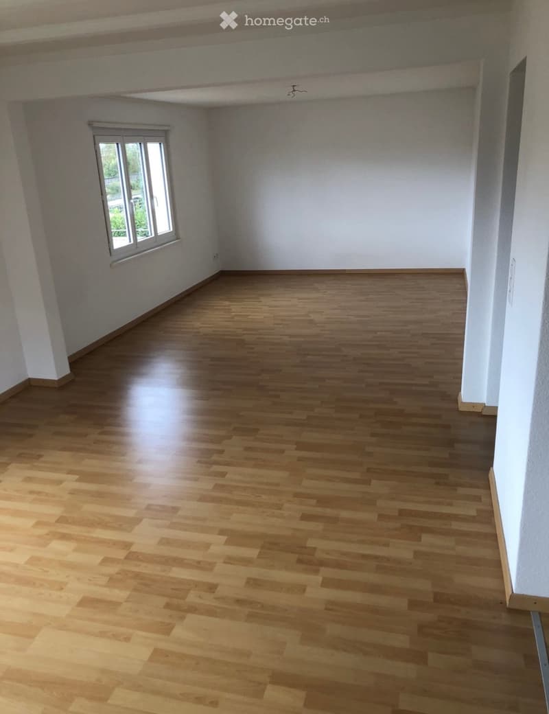 Empty room with wooden flooring and white walls, featuring a single window with white frame and a visible electrical outlet on the wall.