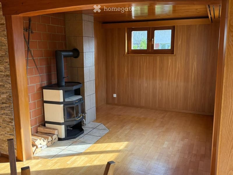Empty room with wooden flooring, exposed wooden beams, brick wall, and a wood-burning stove with a stack of firewood nearby.
