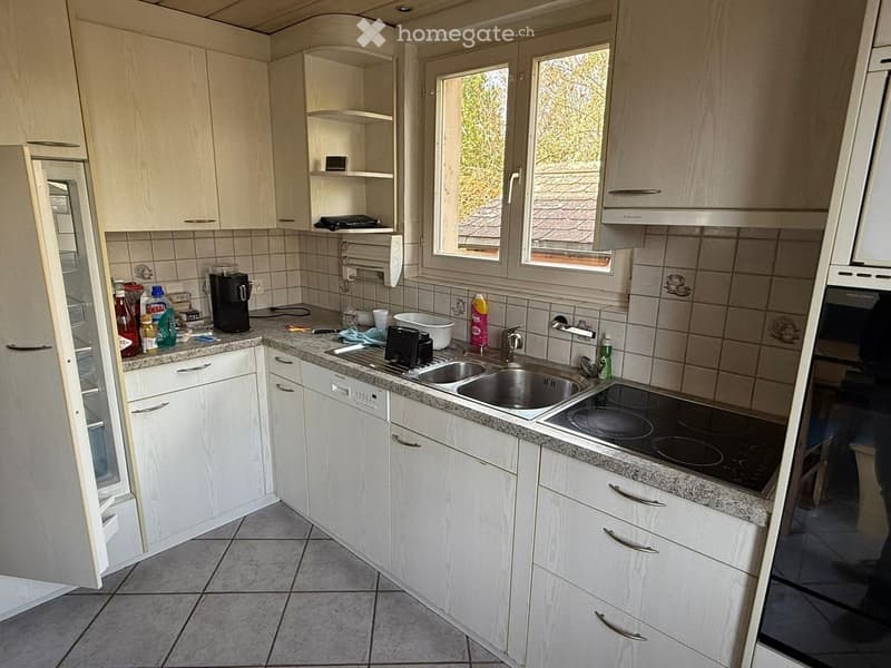 A modern kitchen with white cabinets, granite countertops, a stainless steel sink, and an electric stovetop. The kitchen features a refrigerator, coffee maker, and various cleaning supplies on the counter.