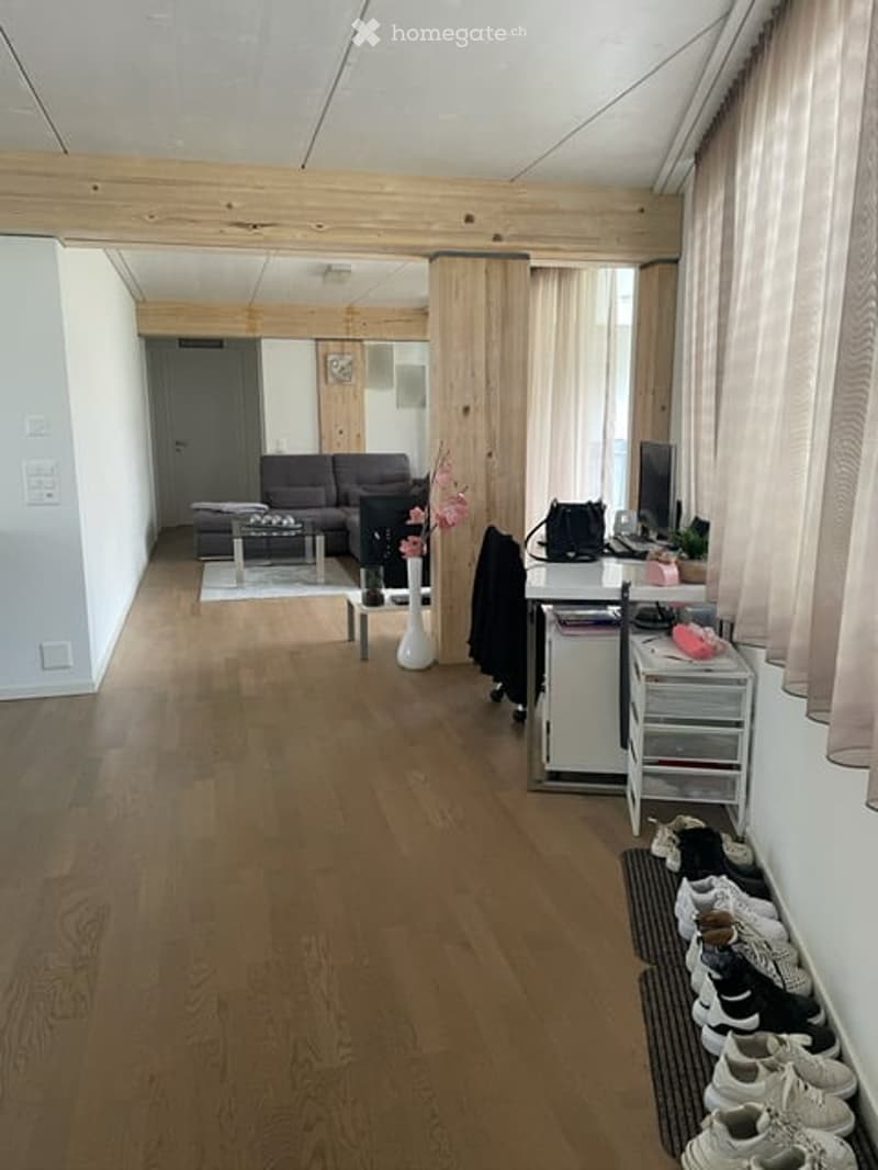 Living room, hardwood floors, white walls, large windows, beige curtains, white vases with pink flowers, gray couch, coffee table, rug, wooden shelves, office desk, monitor, cabinet with drawers, shoes on the floor