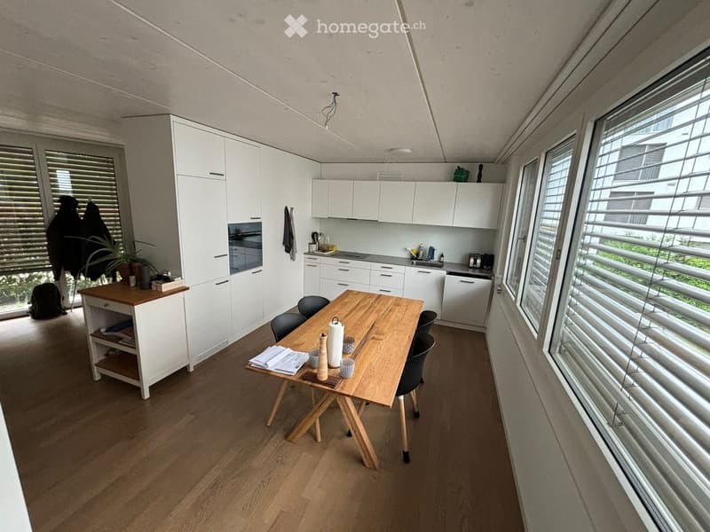 wooden floor, white walls, kitchen with white cabinets, dining table, black chairs, plants, windows with blinds
