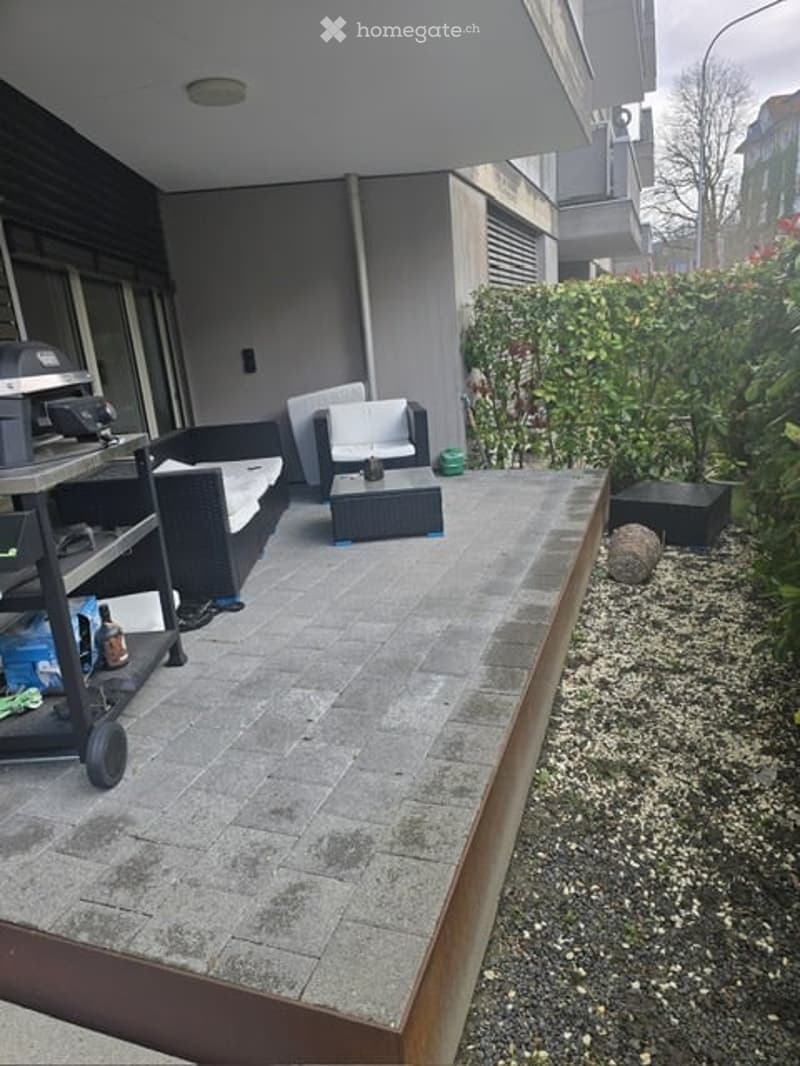 Balcony with outdoor seating area, including a sofa, chairs, and a coffee table, surrounded by greenery and a paved surface.