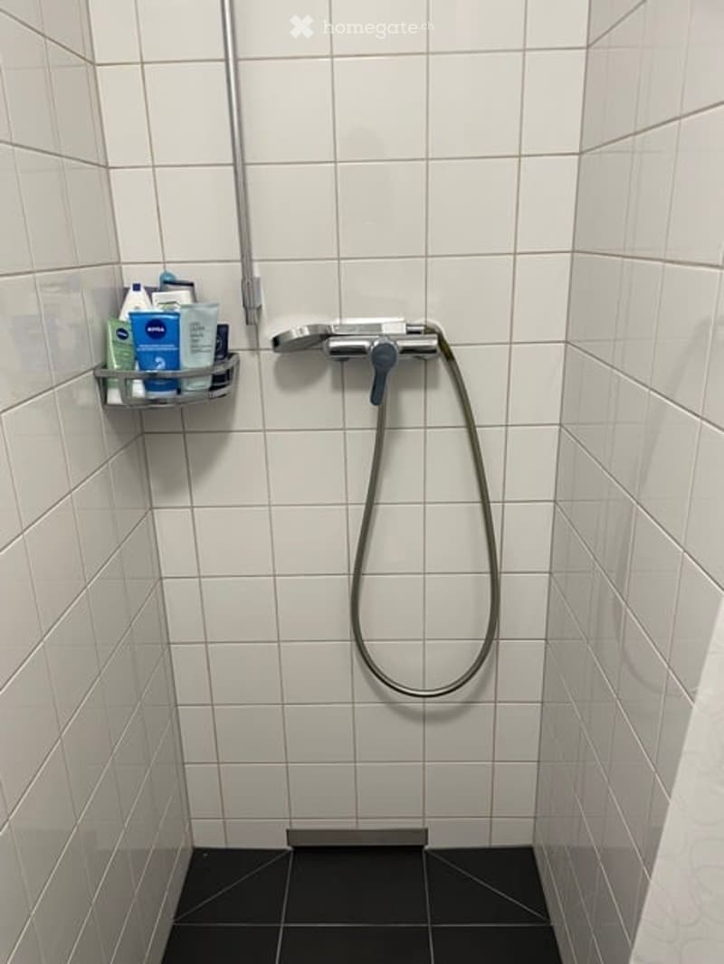 Modern shower stall with white tiled walls, black tiled floor, handheld showerhead, and a small shelf holding toiletries.