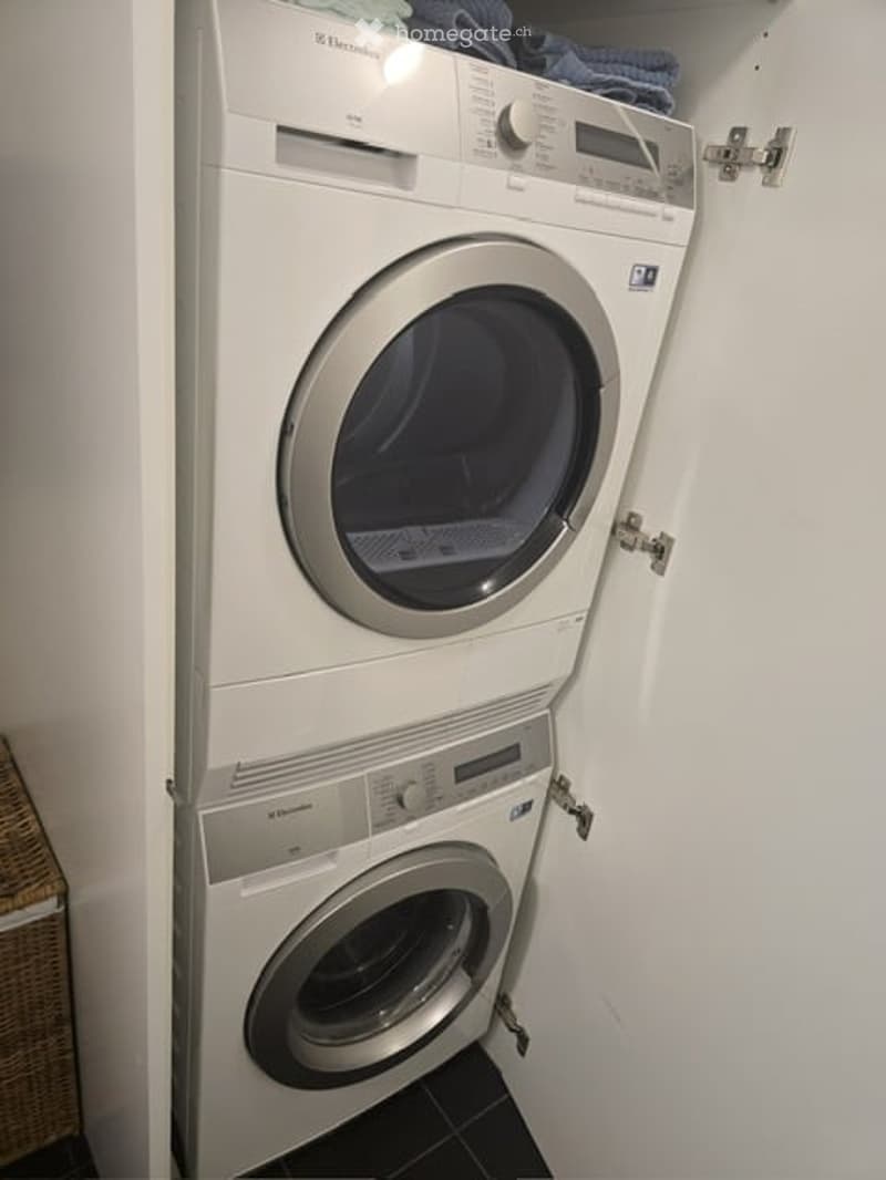 A compact bathroom with a stacked washing machine and dryer unit, white walls, and a small shelf with folded towels on top.