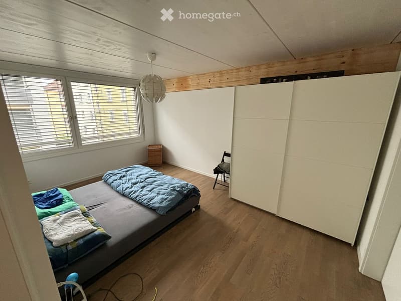 wooden floor, bed with blankets, white closet, chair with clothes, wooden box, large window with blinds, hanging lamp