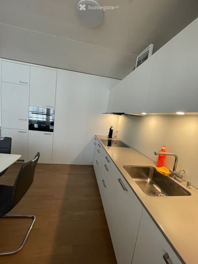 Modern kitchen with wooden floor, built-in appliances, large countertop, white cabinets, modern light fixtures, and modern dining chairs.
