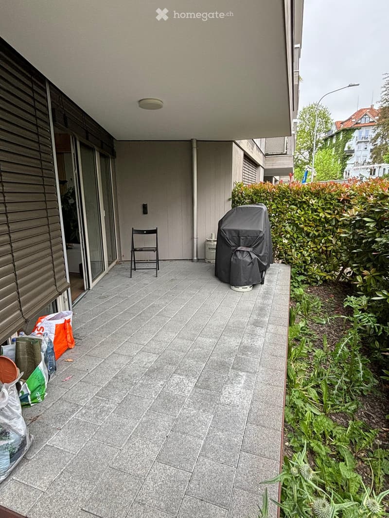 Balcony with tiled floor, closed door with horizontal blinds, table and chair, a gas cylinder, plants, grass