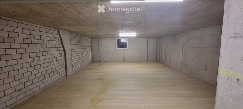 Empty underground parking space with concrete walls, brick sections, and a small window. The floor shows some stains and wear.