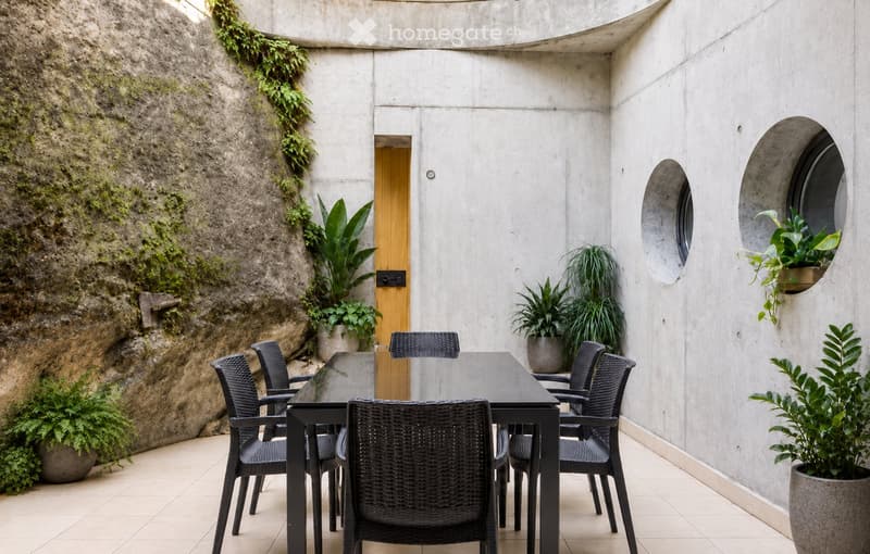 Modern outdoor patio with concrete walls, a dining table with six black wicker chairs, potted plants, and a wooden door leading inside.