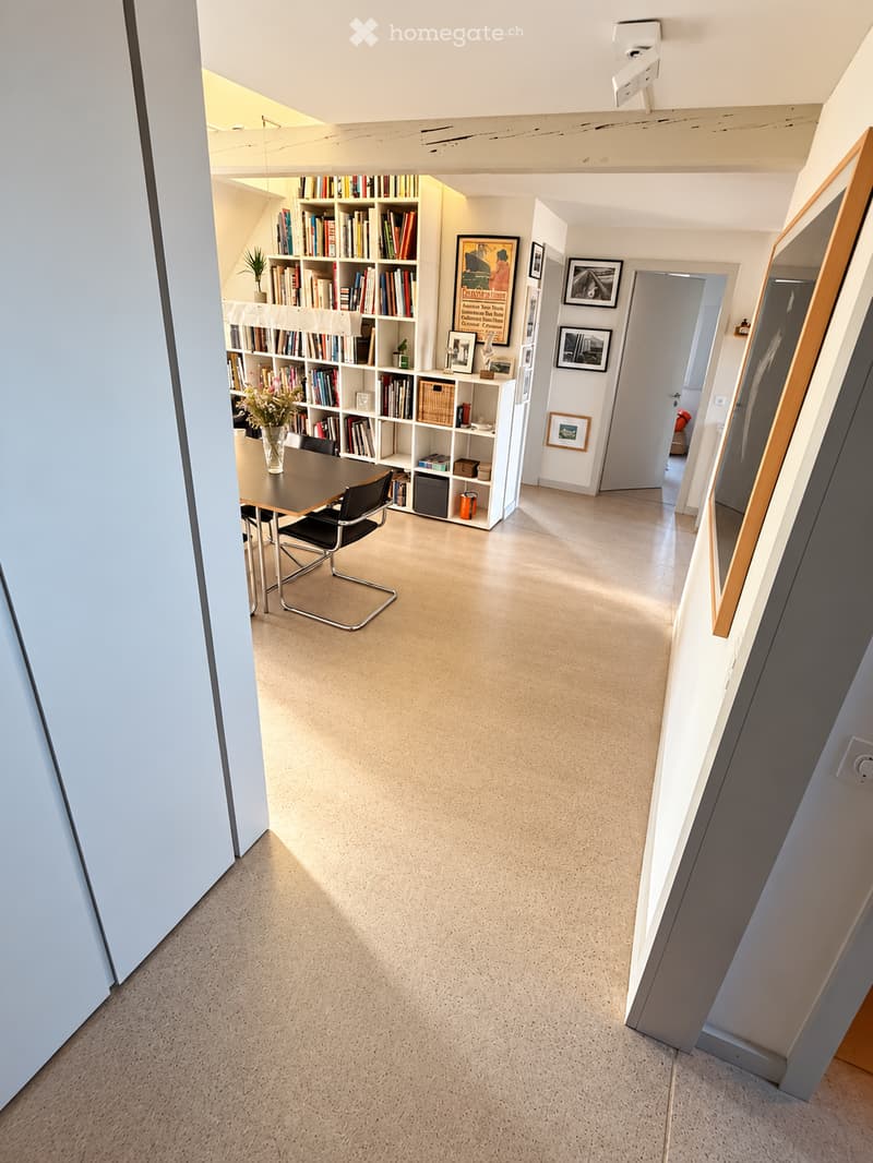 Modern, open-plan room with a bookshelf, dining table, chairs, and decorative items. The space features light-colored walls and a smooth concrete floor.