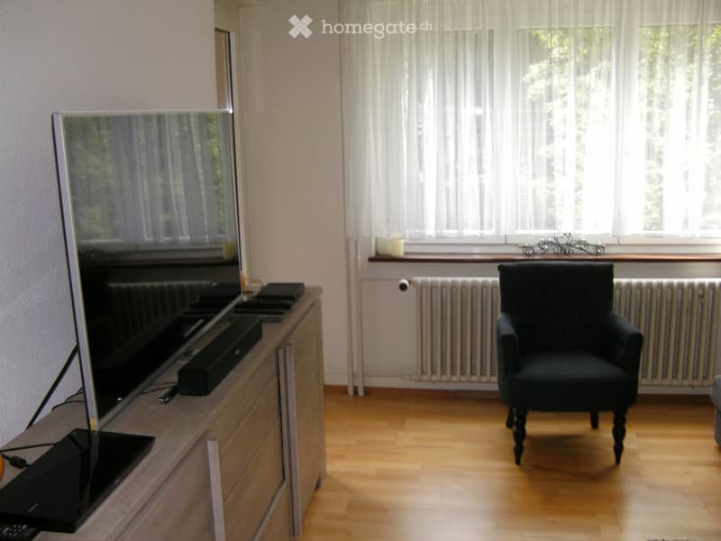 Living room with a large flat-screen TV on a wooden stand, a black armchair, and a window with white curtains.