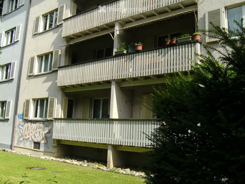 Multi-story residential building with multiple balconies, potted plants, and a grassy area in front.