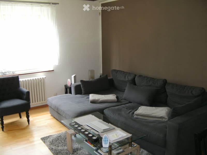 Living room with a dark grey sectional sofa, a glass coffee table, a single armchair, and a small side table. The room has wooden flooring and large windows with white curtains.