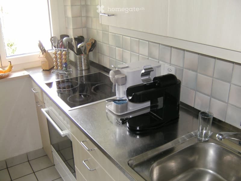 Modern kitchen with white cabinets, stainless steel sink, induction stove, coffee maker, knife block, and various utensils.