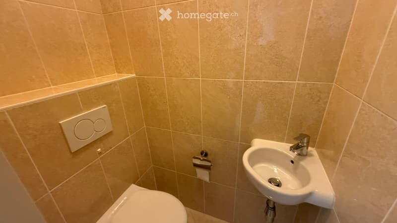 Small bathroom with beige tiled walls and floor, featuring a toilet, a wall-mounted sink with a chrome faucet, and a toilet paper holder.