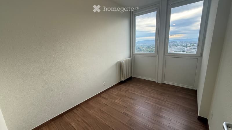 Empty room with wooden flooring, white walls, and large windows offering a scenic view of the city and mountains.