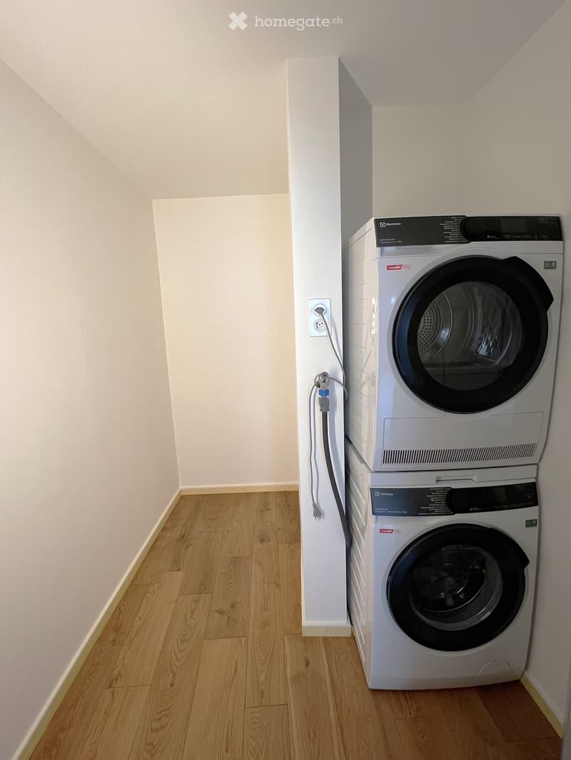 A small, unfurnished room with light wooden flooring and white walls, featuring a stacked washing machine and dryer unit against one wall.