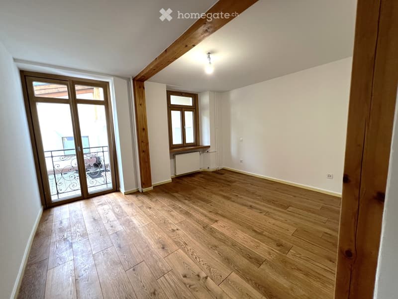 Empty room with wooden flooring, white walls, and large windows, including a sliding glass door leading to a balcony.