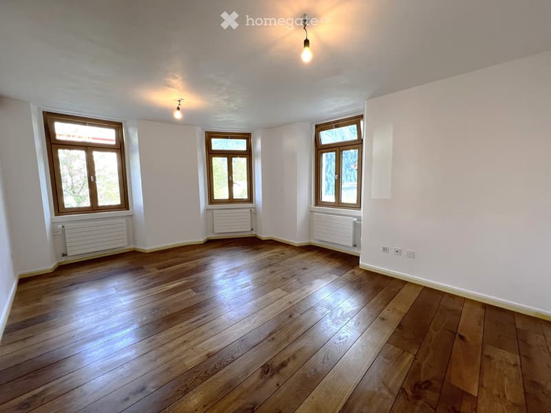 Empty room with wooden flooring, white walls, and three windows with wooden frames. Radiators are present under each window, and the room is illuminated by ceiling lights.