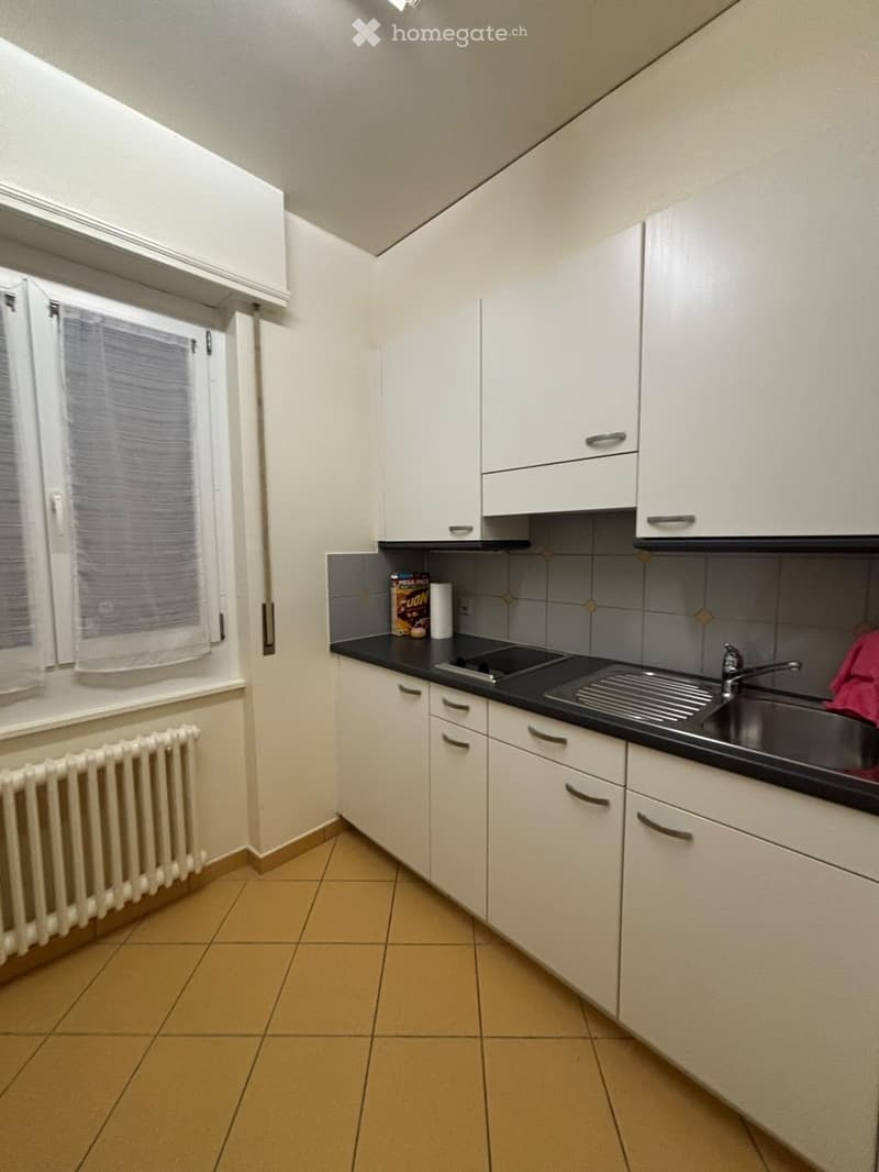 Modern kitchen with white cabinets, black countertop, sink, and window with blinds. Includes a radiator below the window and a few items on the counter.