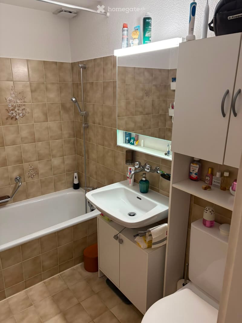 Bathroom with beige tiles, bathtub, sink, toilet, and various toiletries on shelves and counters.
