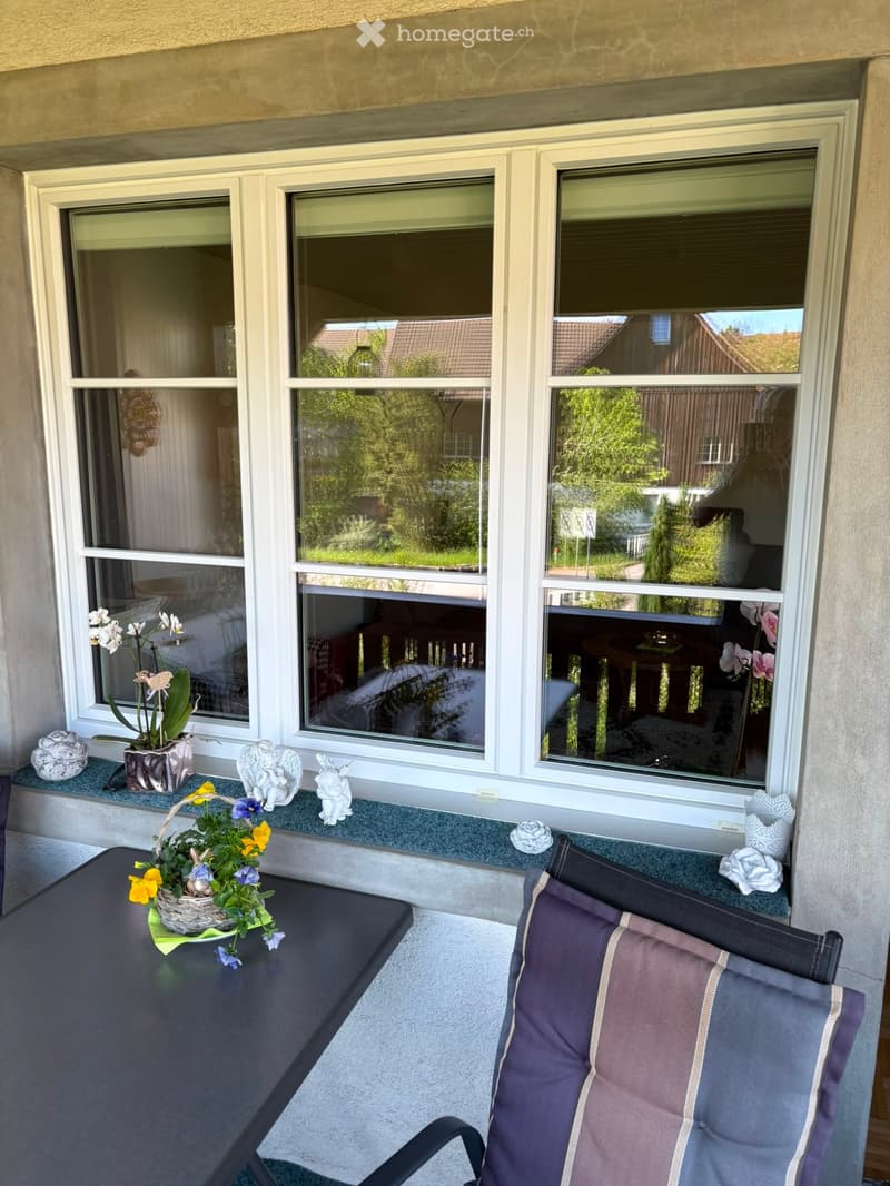 Balcony with large windows, a black table with a flower arrangement, and a padded chair with cushions.