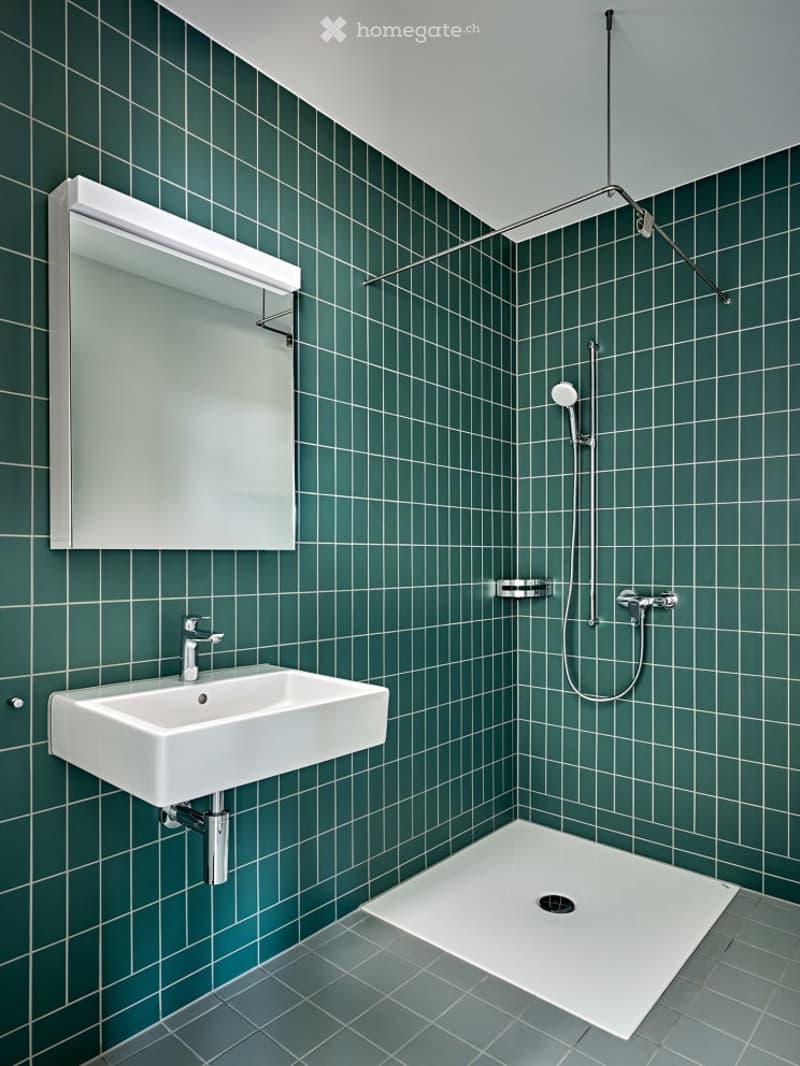 Modern bathroom with green tiled walls, white sink, mirror, and shower area.