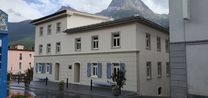 Two-story building with light gray facade, multiple windows, blue shutters, and a view of mountains in the background.